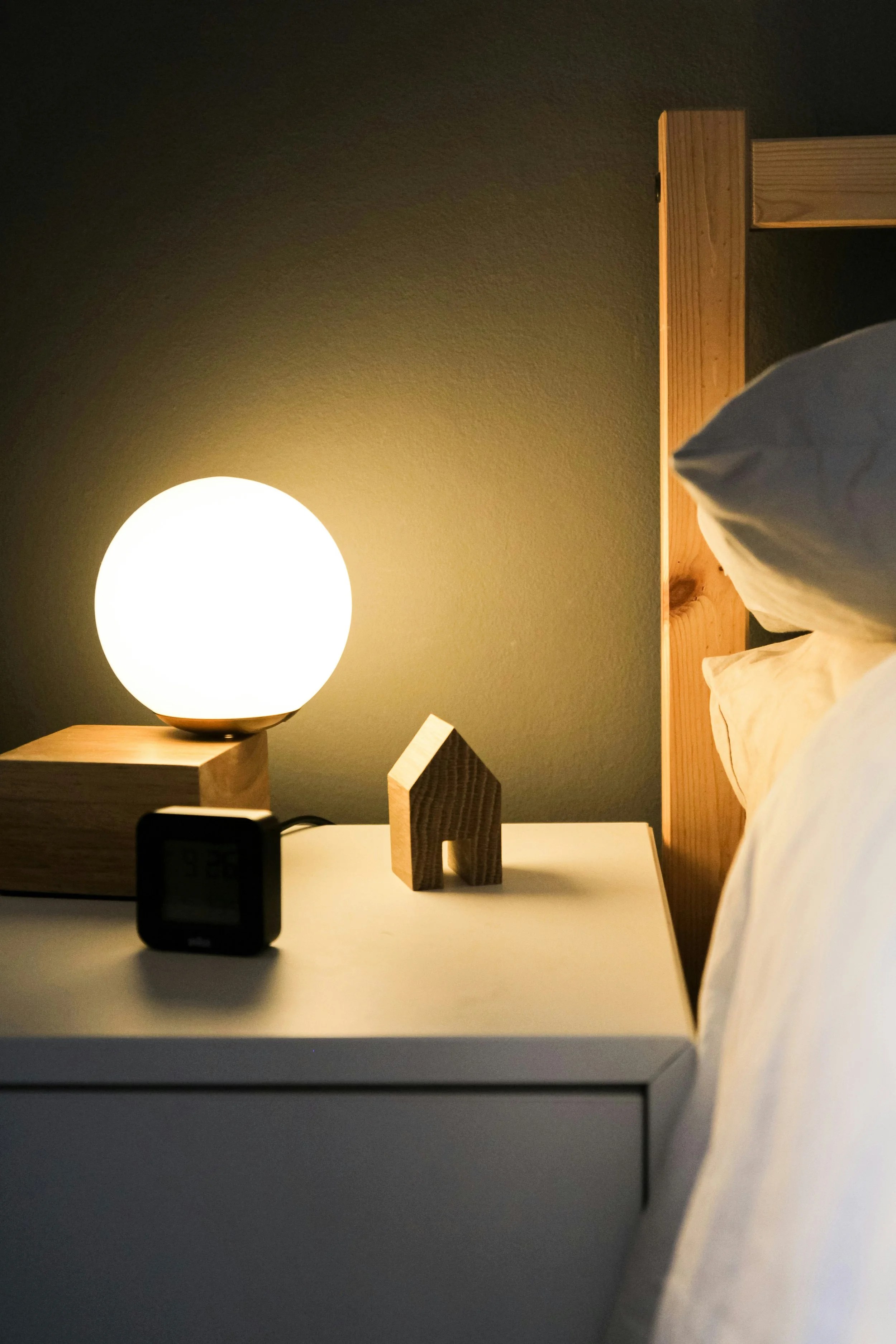 A bedside table featuring a round lamp, a small wooden house figurine, and a digital clock, with a bed and pillow partially visible in the background.