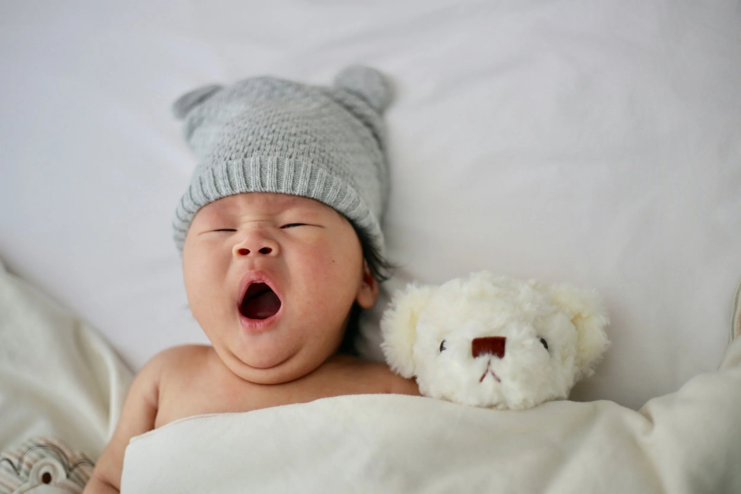 A baby yawning while wearing a gray knit hat, next to a white stuffed bear, resting on a soft blanket.