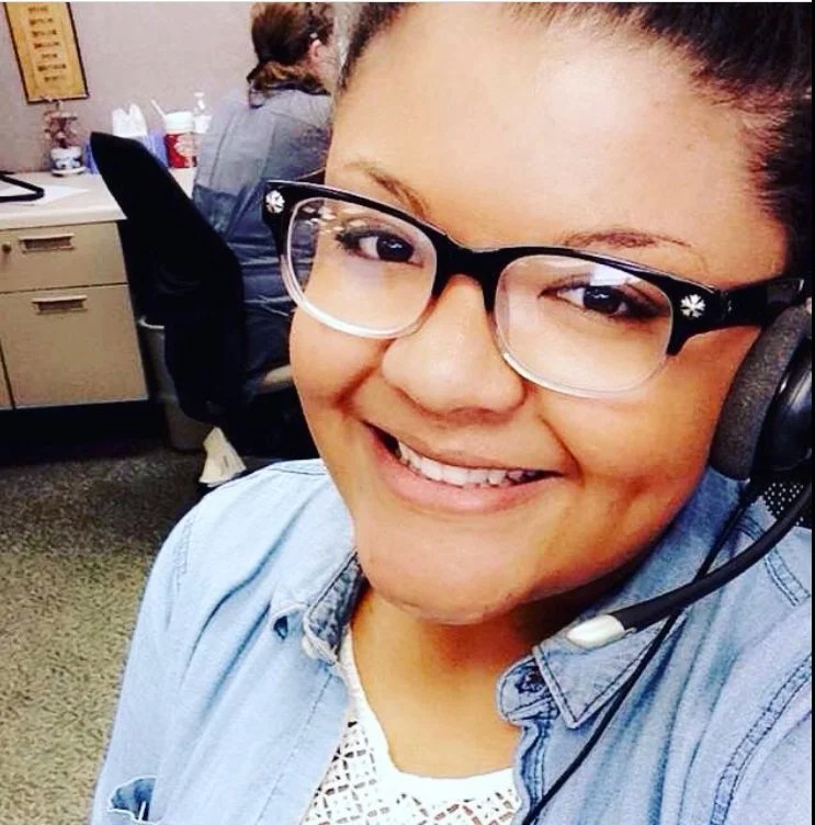 A smiling woman wearing glasses and a headset, posing for a selfie in an office setting with a coworker visible in the background.