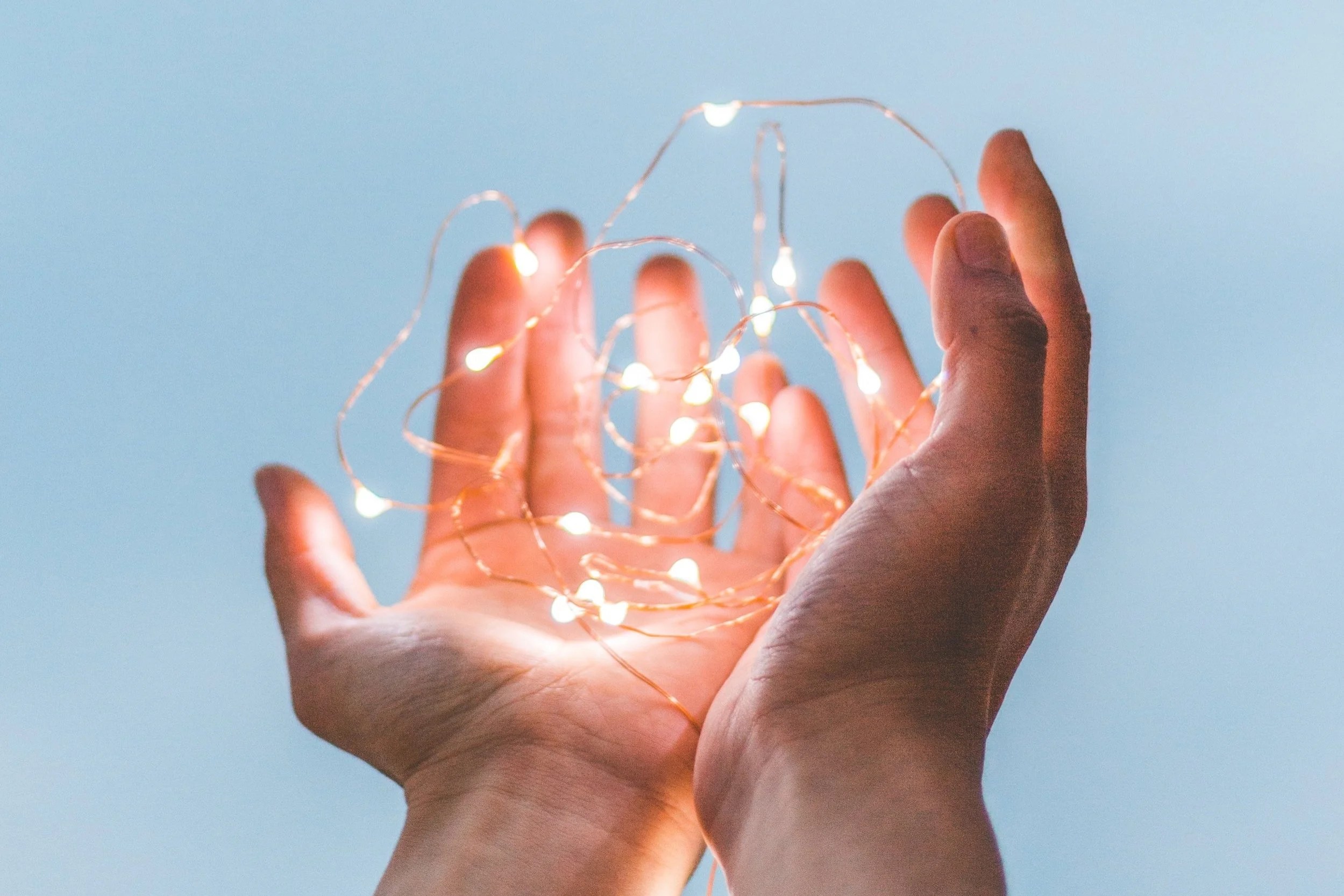 Two hands holding a tangled string of warm white fairy lights against a soft blue background.