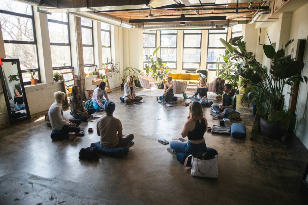 A group of people sitting in a circle on a floor in a well-lit room with large windows and plants, engaged in a mindfulness or meditation session.
