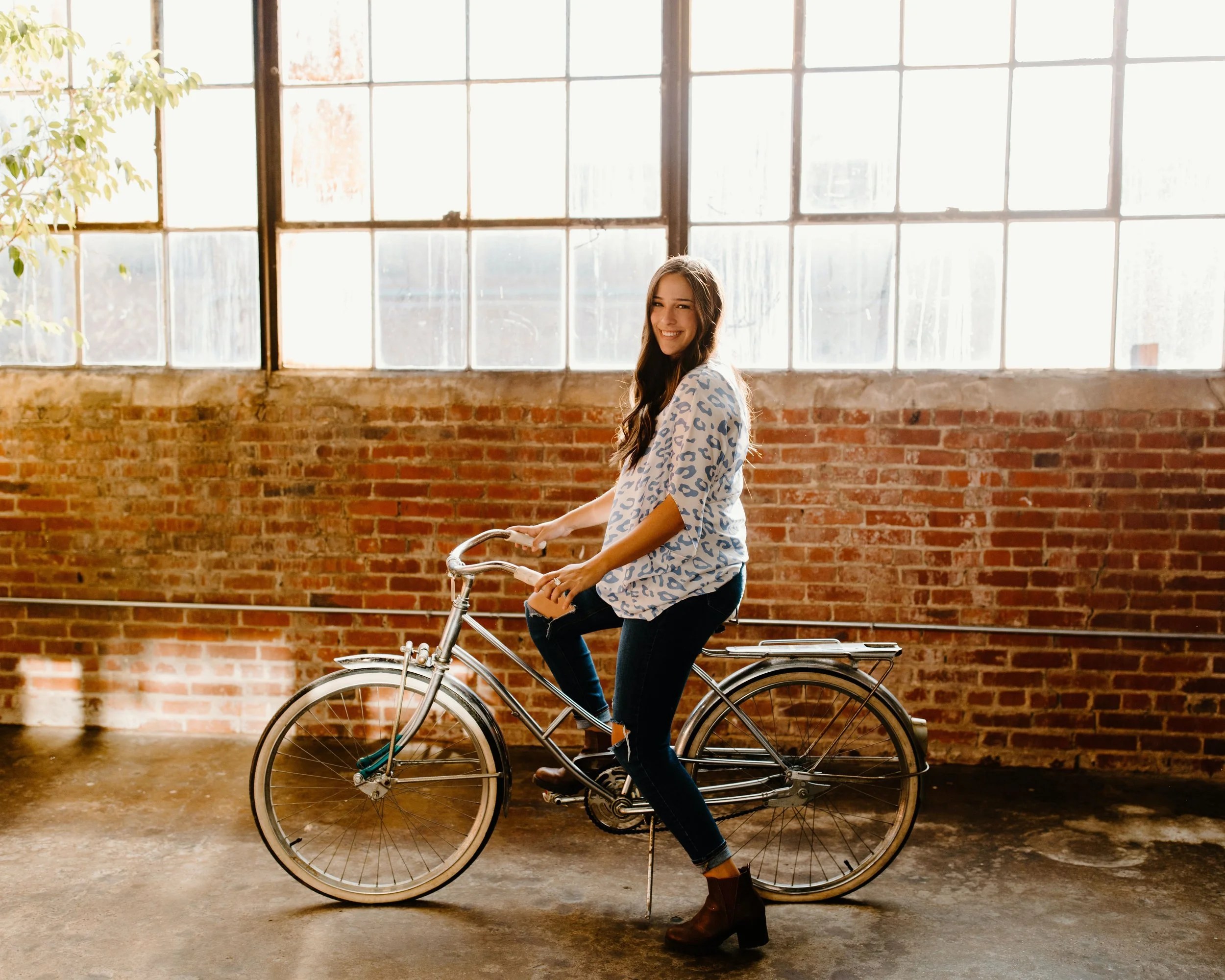 A young woman sitting on a vintage bicycle inside a warehouse with large windows and exposed brick walls, smiling at the camera.
