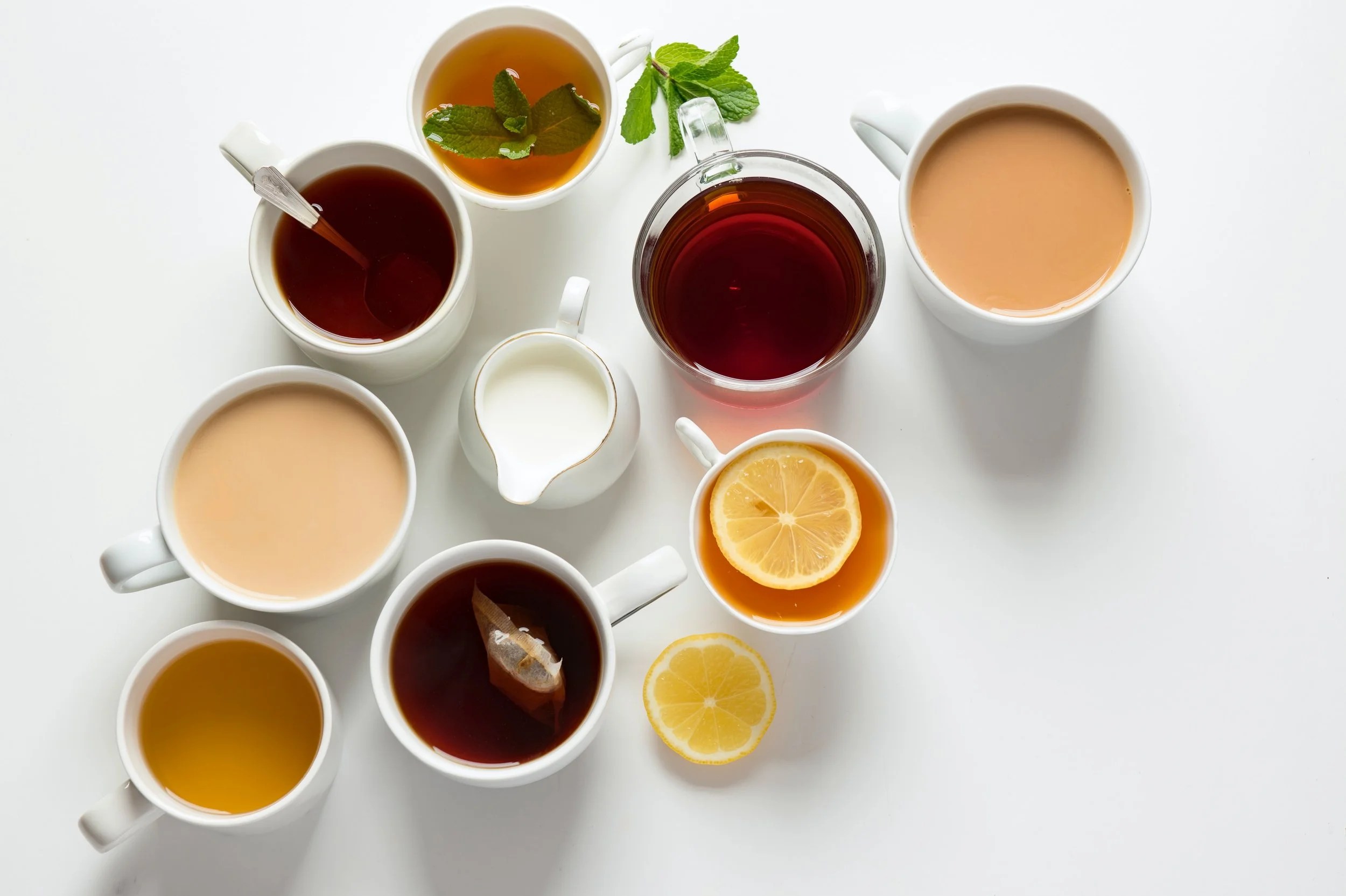 An assortment of tea cups featuring different types of tea, including black tea, green tea, and herbal tea, with a lemon slice and mint leaves for garnish on a white surface.