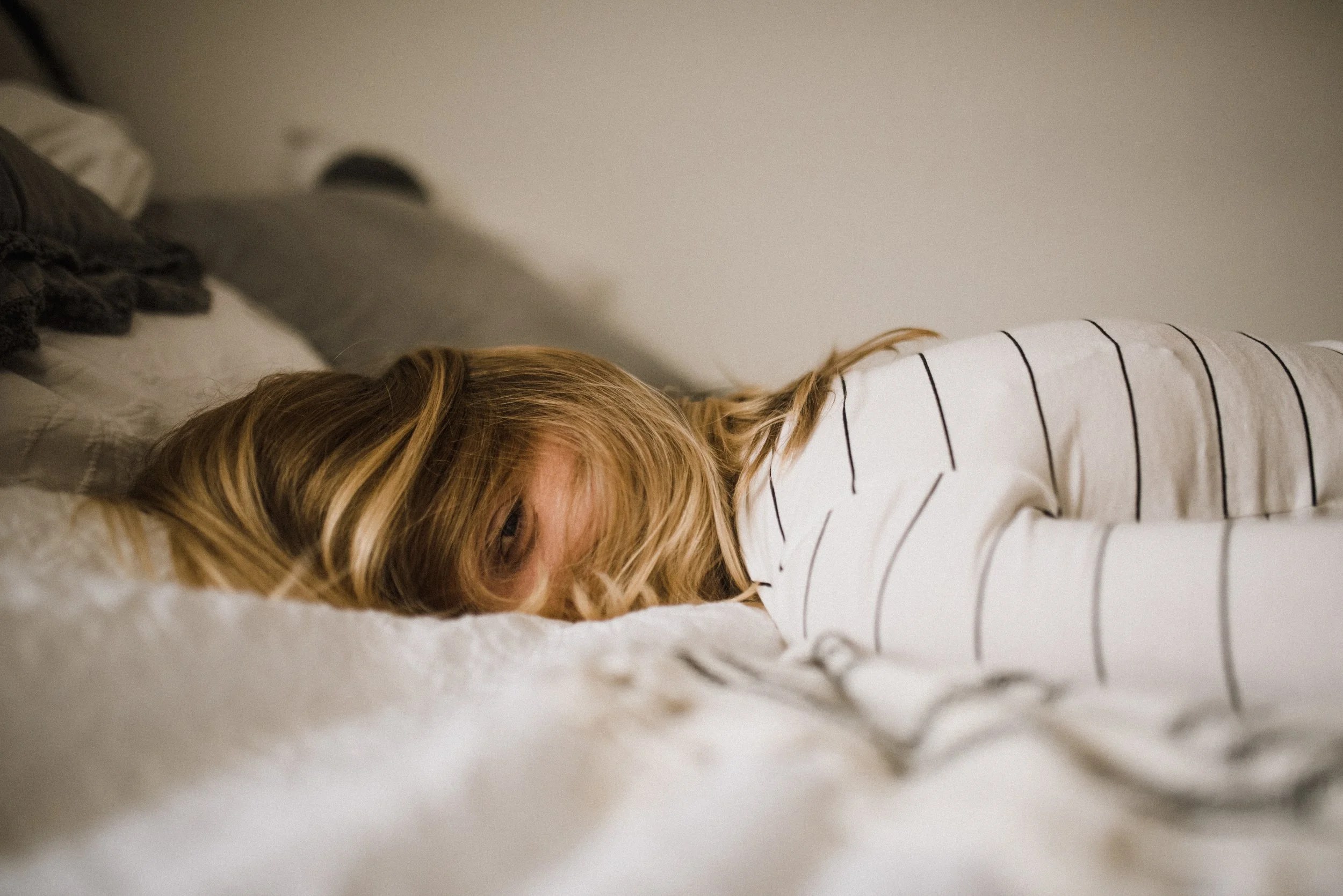 A person lying face down on a bed, partially obscured by their hair, wearing a white striped long-sleeve shirt.