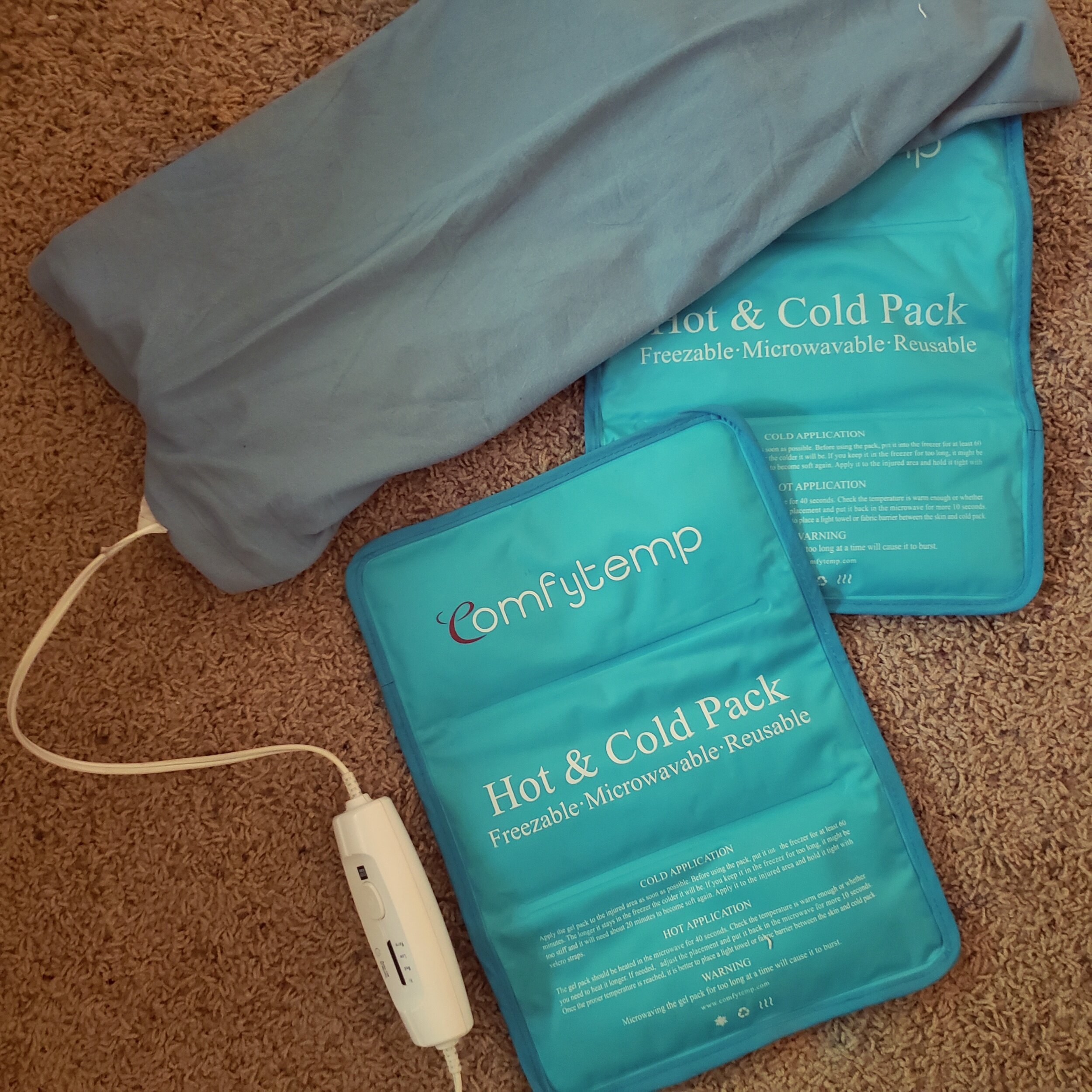 A blue heating pad with a connected control unit and two reusable hot and cold packs on a beige carpet.