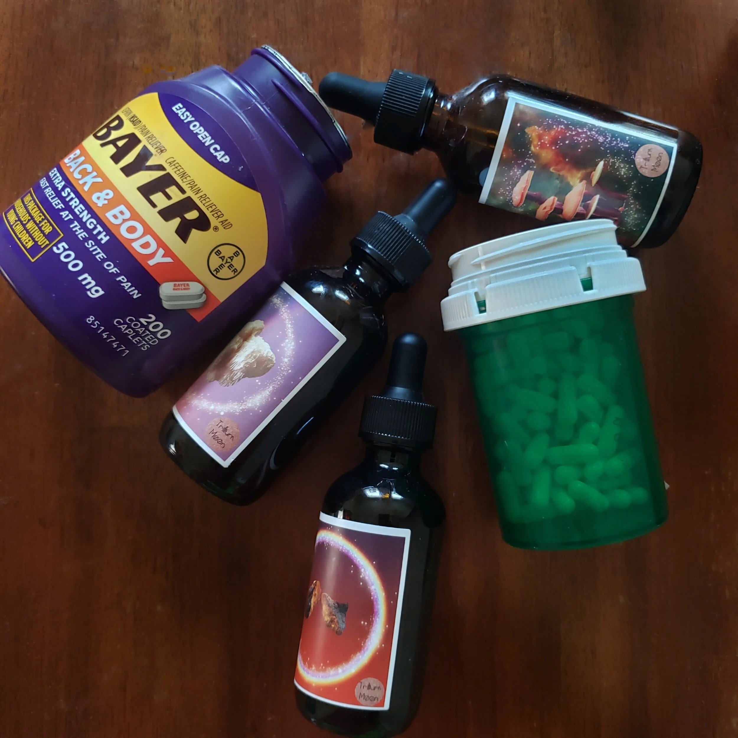A variety of health products including Bayer Back & Body pain relief, three dropper bottles with colorful labels, and a green prescription container filled with green capsules, arranged on a wooden surface.