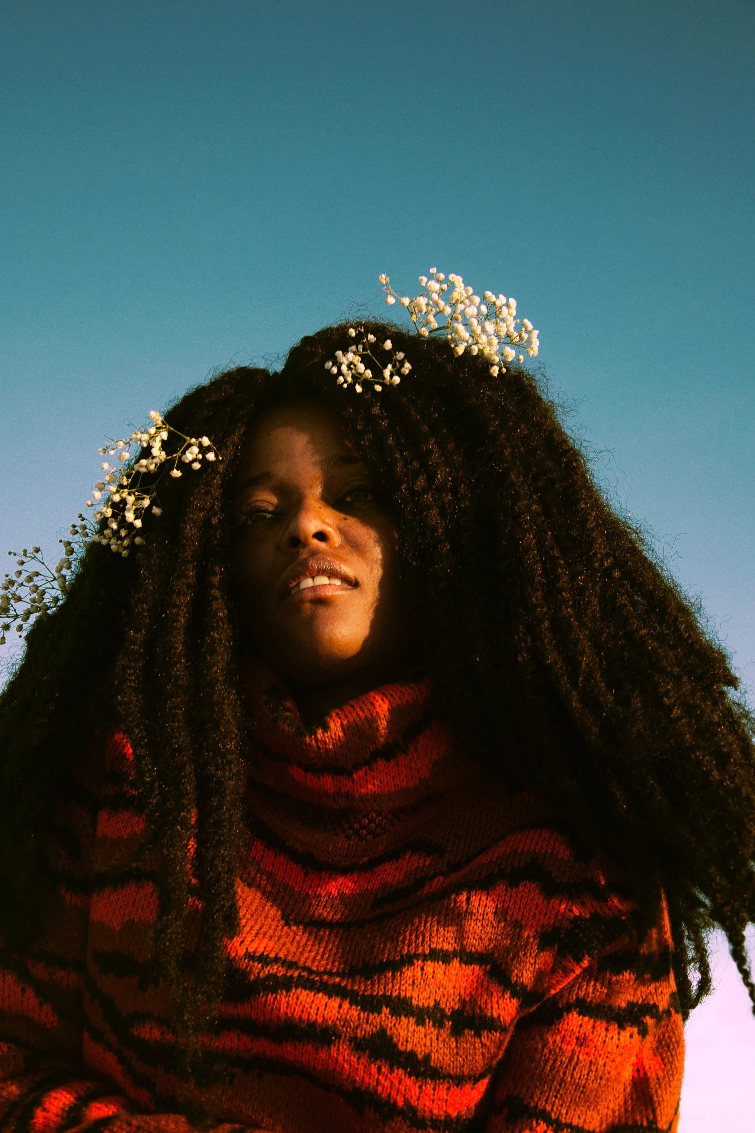 A young woman with long, curly hair adorned with white flowers, wearing a red and black striped sweater, gazes thoughtfully against a clear blue sky.