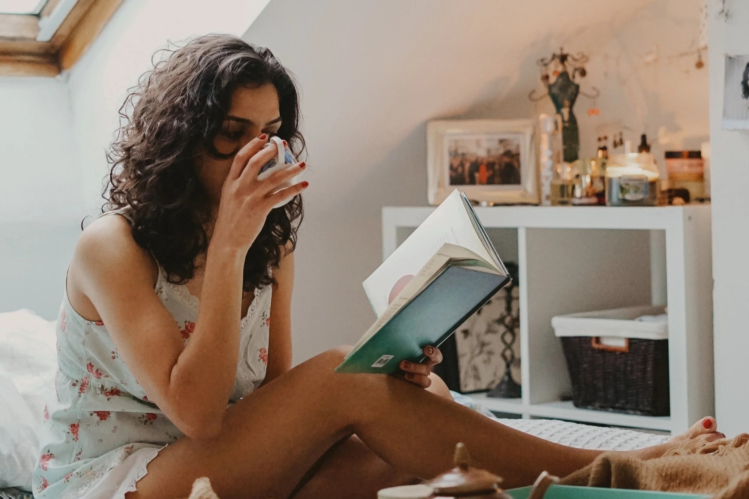 A woman sits on a bed, reading a book while holding a cup. She has curly hair and is wearing a floral nightgown. In the background, there are decorative items and a window.