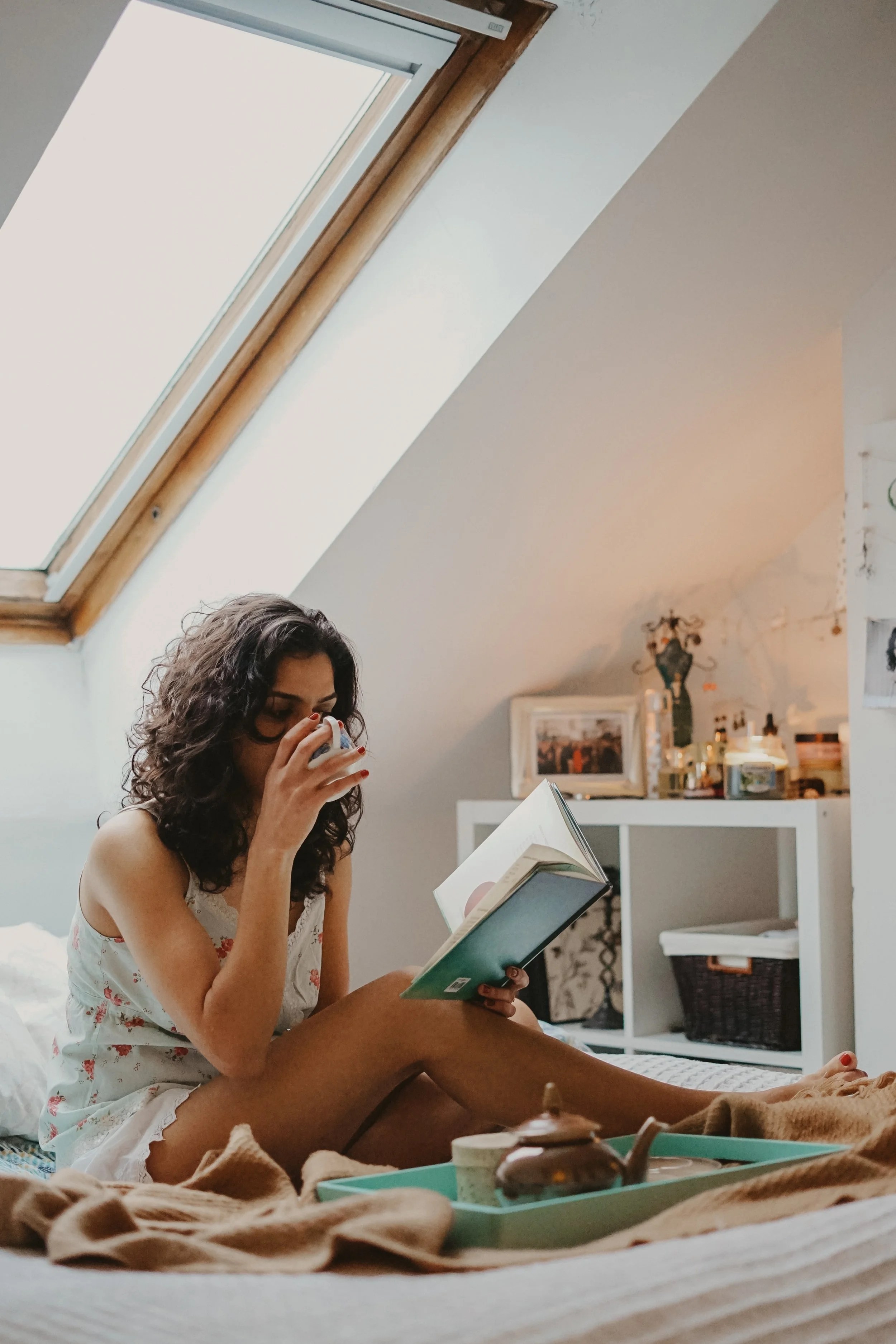 A woman with curly hair wearing a floral-patterned sleepwear sits on a bed, sipping from a white cup while reading a book in a cozy room with a skylight.